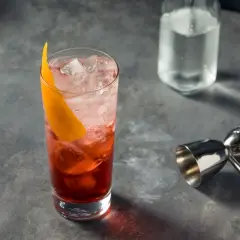 Un verre rempli de glaçons et d'un cocktail rouge vif, garni d'une écorce d'orange. Un jigger et une bouteille d'eau gazeuse sont à proximité, prêts pour un martini rosso.