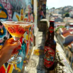 Une main tient un verre à martini pétillant, à côté d'une bouteille de bière, sur fond de graffitis colorés et d'une ville ensoleillée.