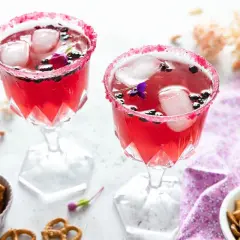 Deux verres de **meilleur cocktail mousseux** rose vif, décorés de fruits rouges et de fleurs, avec des bretzels et des noix en accompagnement.