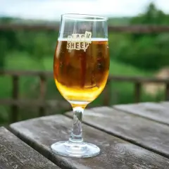Un verre de bière ale blonde, marqué "Black Sheep", sur une table en bois avec un fond verdoyant.