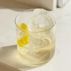 Un cocktail vermouth rafraîchissant avec une grosse boule de glace et un zeste de citron sur un comptoir blanc.