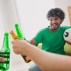 Un homme souriant, ballon de foot sous le bras, trinque avec des bières après le sport.