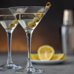 Deux verres à martini remplis, garni d'olives. Une recette de dirty martini à portée de main, avec des citrons et un shaker en arrière-plan.