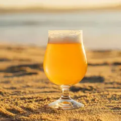 Un verre de bière blonde, type IPA, sur le sable chaud, avec la mer en arrière-plan au coucher du soleil.