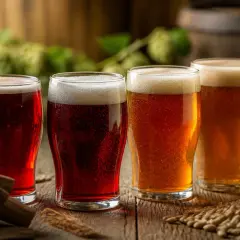 Quatre verres de bière, dont deux bières rouges foncées, sur une table en bois avec du houblon et du malt en arrière-plan.