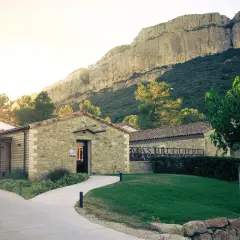 Le Mas Bruguière, un domaine viticole en pierre, se dresse au pied d'une falaise imposante, baigné par la lumière dorée du soleil couchant.