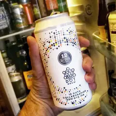 Une main tient une bière canette forte "12 Acres Dot Brew" devant un réfrigérateur rempli de bières.