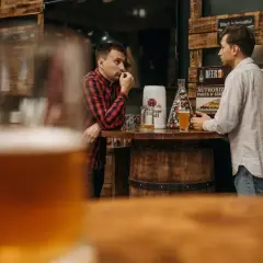 Deux hommes discutent près d'un bar en bois, une bière la bête à la main, dans une ambiance rustique.