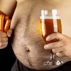 Un homme tient un verre de bière et des saucisses, soulevant la question : la bière fait elle grossir ? Son ventre proéminent suggère une réponse.