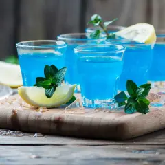 Cocktail martini blanc curaçao, bleu vif, garni de menthe fraîche et de quartiers de citron, sur un plateau en bois avec de la glace pilée.
