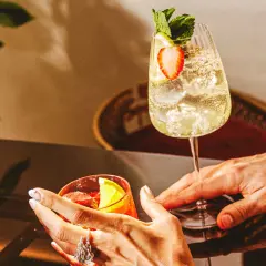 Deux mains tendent des cocktails rafraîchissants, un verre de martini bitter rouge et un autre pétillant garni de fraises et de menthe.