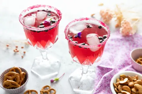 Deux verres de **meilleur cocktail mousseux** rose vif, décorés de fruits rouges et de fleurs, avec des bretzels et des noix en accompagnement.