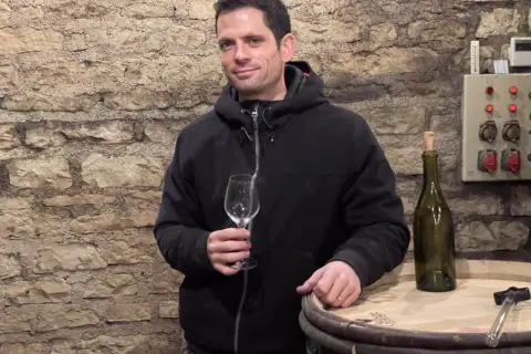 Un homme tient un verre vide dans une cave à vin, à côté d'une bouteille et d'un tonneau. Peut-être une dégustation de Romanée-Conti ?