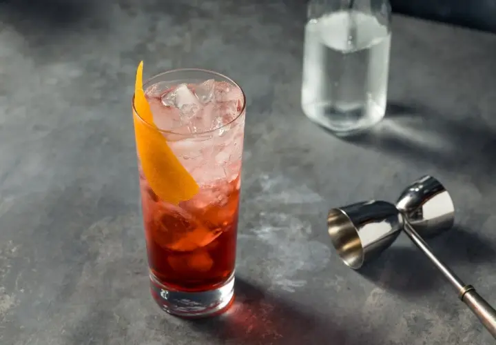 Un verre rempli de glaçons et d'un cocktail rouge vif, garni d'une écorce d'orange. Un jigger et une bouteille d'eau gazeuse sont à proximité, prêts pour un martini rosso.