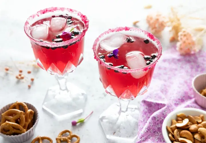 Deux verres de **meilleur cocktail mousseux** rose vif, décorés de fruits rouges et de fleurs, avec des bretzels et des noix en accompagnement.