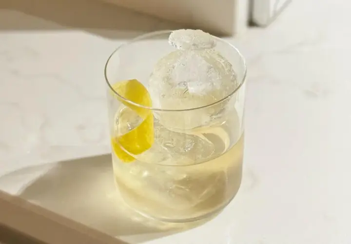 Un cocktail vermouth rafraîchissant avec une grosse boule de glace et un zeste de citron sur un comptoir blanc.