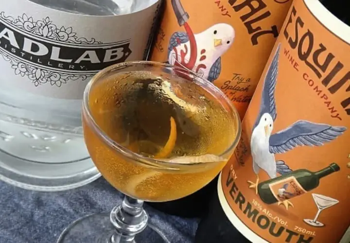 Un cocktail martini doré, garni d'un zeste d'orange, est prêt à être dégusté, entouré de bouteilles de gin et de vermouth.