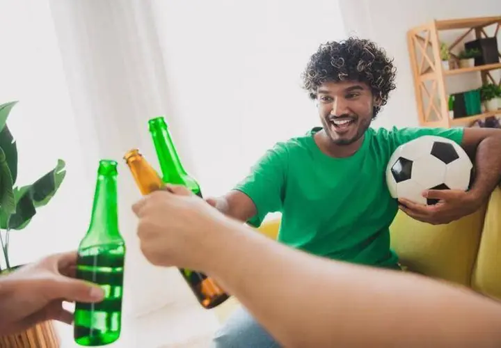 Un homme souriant, ballon de foot sous le bras, trinque avec des bières après le sport.