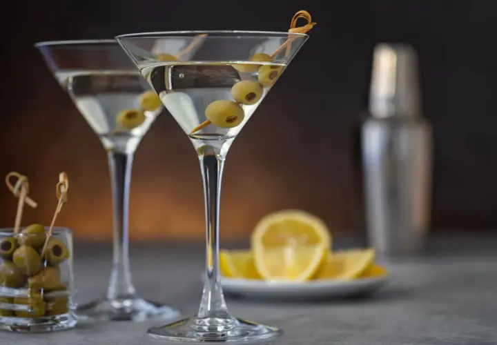 Deux verres à martini remplis, garni d'olives. Une recette de dirty martini à portée de main, avec des citrons et un shaker en arrière-plan.