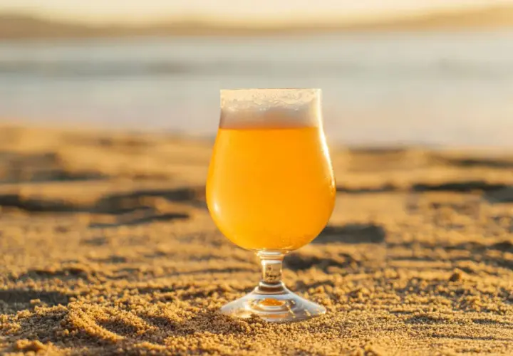 Un verre de bière blonde, type IPA, sur le sable chaud, avec la mer en arrière-plan au coucher du soleil.