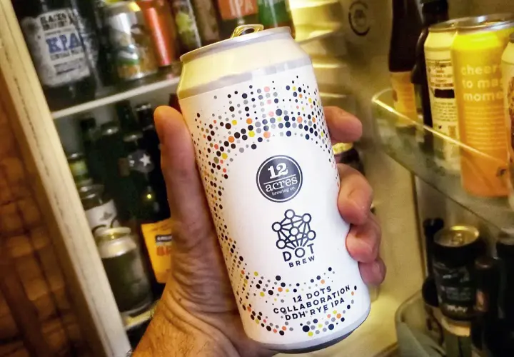 Une main tient une bière canette forte "12 Acres Dot Brew" devant un réfrigérateur rempli de bières.