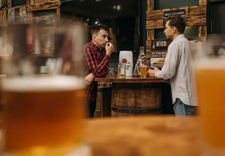 Deux hommes discutent près d'un bar en bois, une bière la bête à la main, dans une ambiance rustique.