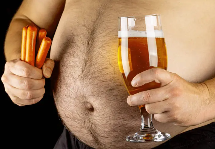 Un homme tient un verre de bière et des saucisses, soulevant la question : la bière fait elle grossir ? Son ventre proéminent suggère une réponse.