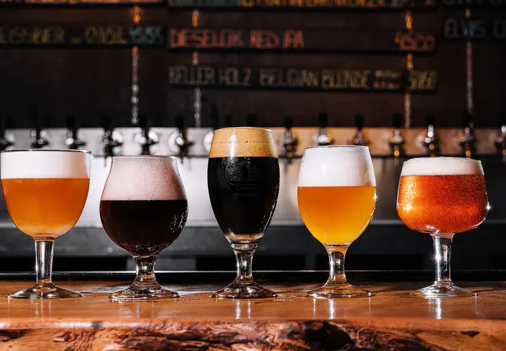 Cinq verres de bière, dont une pale ale ou ipa, sur un comptoir en bois. Derrière, des tireuses et un tableau des bières.