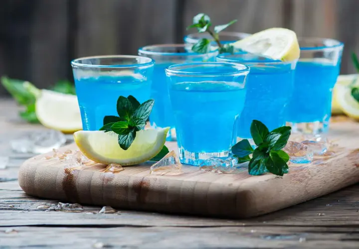 Cocktail martini blanc curaçao, bleu vif, garni de menthe fraîche et de quartiers de citron, sur un plateau en bois avec de la glace pilée.