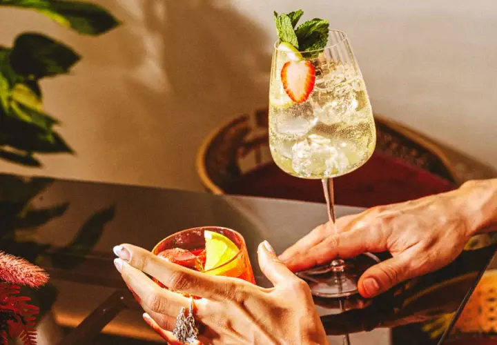 Deux mains tendent des cocktails rafraîchissants, un verre de martini bitter rouge et un autre pétillant garni de fraises et de menthe.