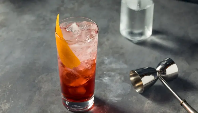 Un verre rempli de glaçons et d'un cocktail rouge vif, garni d'une écorce d'orange. Un jigger et une bouteille d'eau gazeuse sont à proximité, prêts pour un martini rosso.