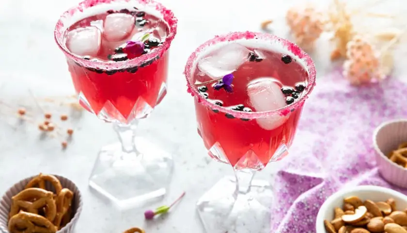 Deux verres de **meilleur cocktail mousseux** rose vif, décorés de fruits rouges et de fleurs, avec des bretzels et des noix en accompagnement.