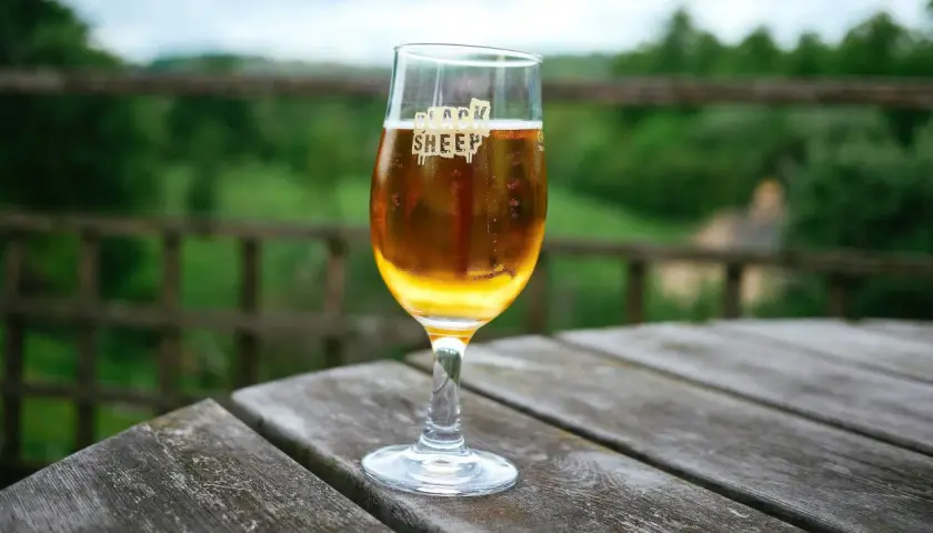 Un verre de bière ale blonde, marqué "Black Sheep", sur une table en bois avec un fond verdoyant.