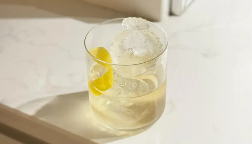 Un cocktail vermouth rafraîchissant avec une grosse boule de glace et un zeste de citron sur un comptoir blanc.