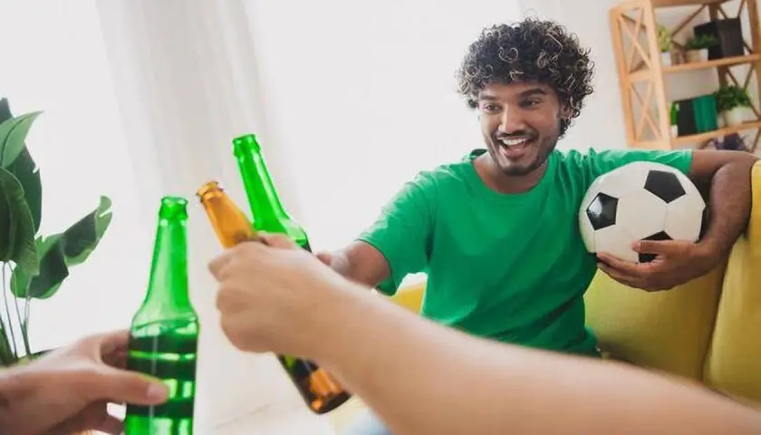 Un homme souriant, ballon de foot sous le bras, trinque avec des bières après le sport.