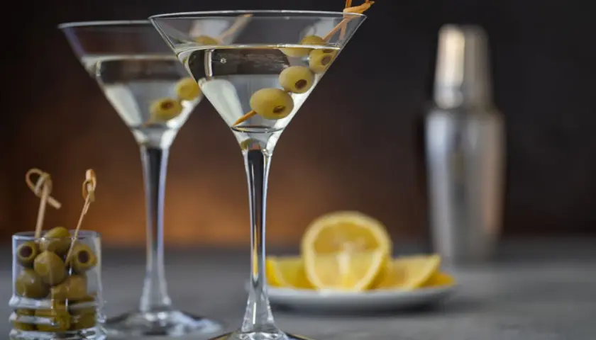 Deux verres à martini remplis, garni d'olives. Une recette de dirty martini à portée de main, avec des citrons et un shaker en arrière-plan.