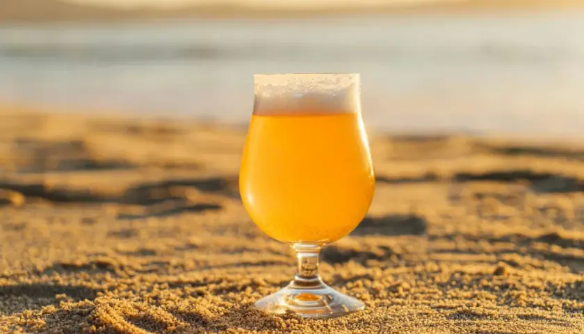 Un verre de bière blonde, type IPA, sur le sable chaud, avec la mer en arrière-plan au coucher du soleil.