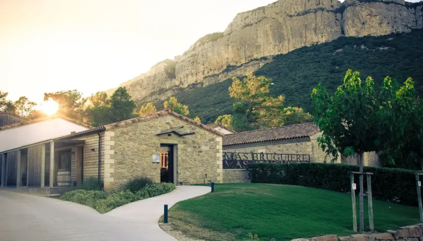 Le Mas Bruguière, un domaine viticole en pierre, se dresse au pied d'une falaise imposante, baigné par la lumière dorée du soleil couchant.