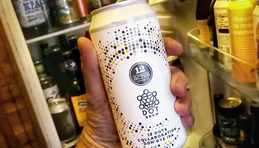 Une main tient une bière canette forte "12 Acres Dot Brew" devant un réfrigérateur rempli de bières.