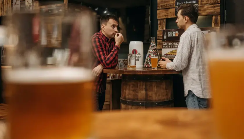 Deux hommes discutent près d'un bar en bois, une bière la bête à la main, dans une ambiance rustique.