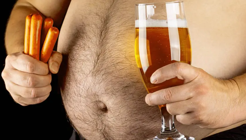Un homme tient un verre de bière et des saucisses, soulevant la question : la bière fait elle grossir ? Son ventre proéminent suggère une réponse.