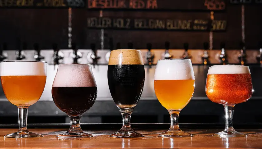 Cinq verres de bière, dont une pale ale ou ipa, sur un comptoir en bois. Derrière, des tireuses et un tableau des bières.