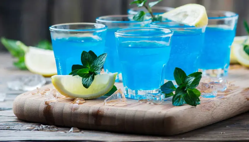 Cocktail martini blanc curaçao, bleu vif, garni de menthe fraîche et de quartiers de citron, sur un plateau en bois avec de la glace pilée.