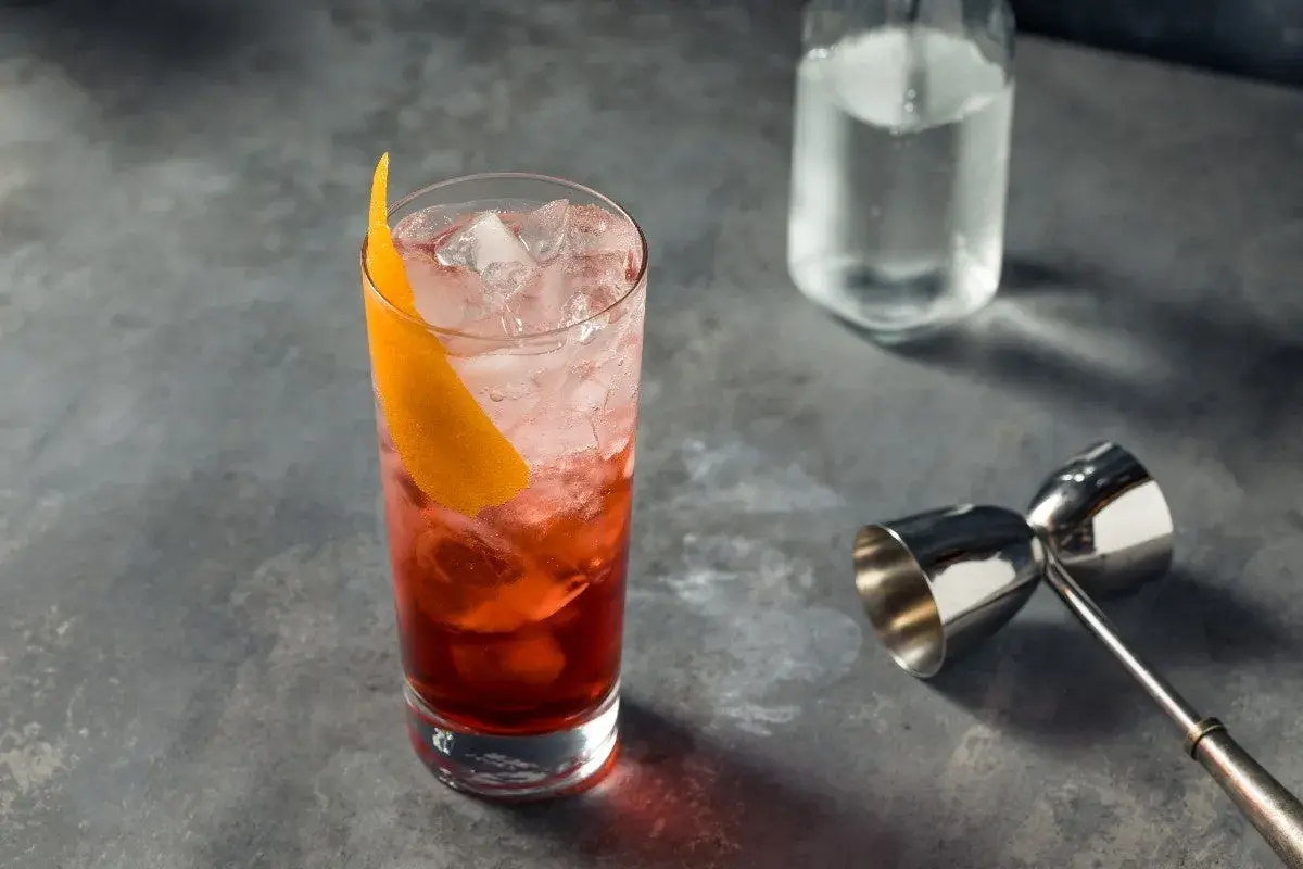 Un verre rempli de glaçons et d'un cocktail rouge vif, garni d'une écorce d'orange. Un jigger et une bouteille d'eau gazeuse sont à proximité, prêts pour un martini rosso.