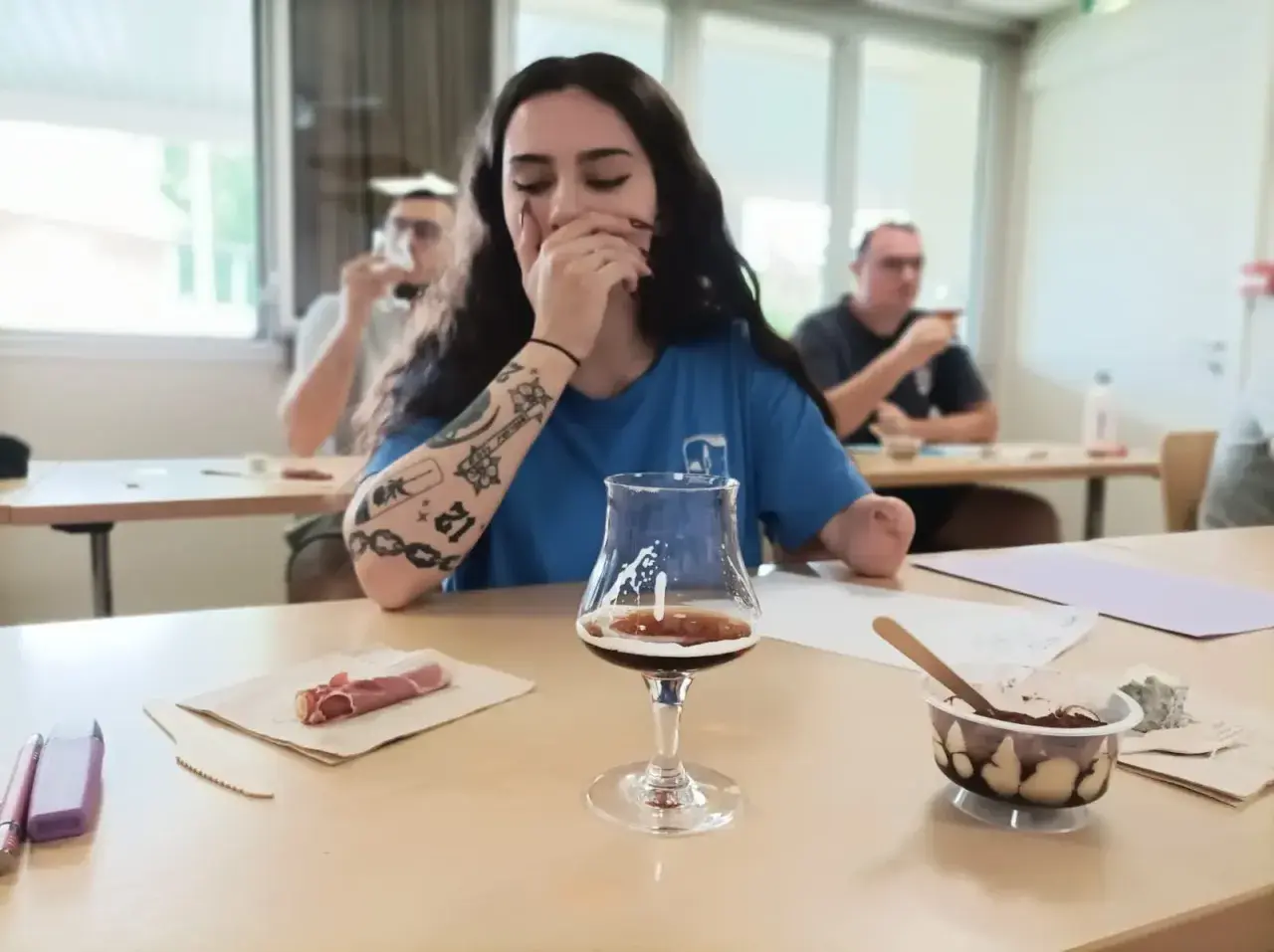 Jeune femme aux tatouages participe à une formation vin, dégustant une bière artisanale avec des accompagnements.