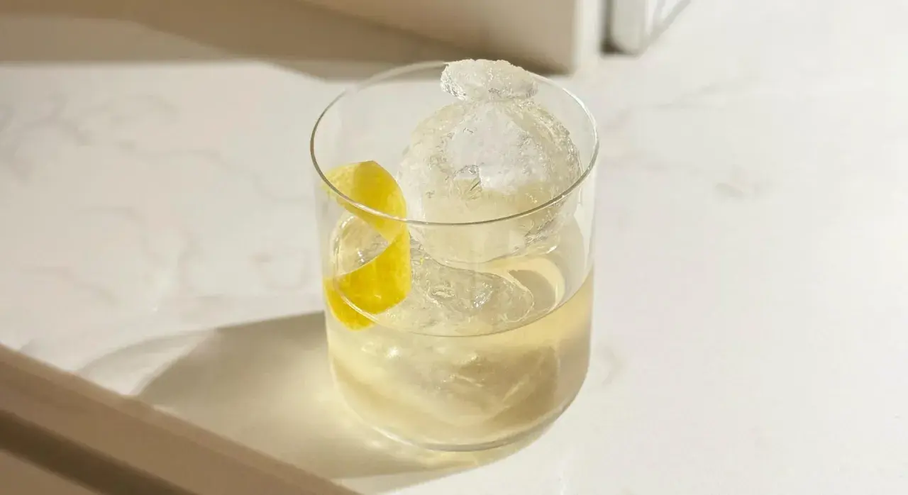 Un cocktail vermouth rafraîchissant avec une grosse boule de glace et un zeste de citron sur un comptoir blanc.