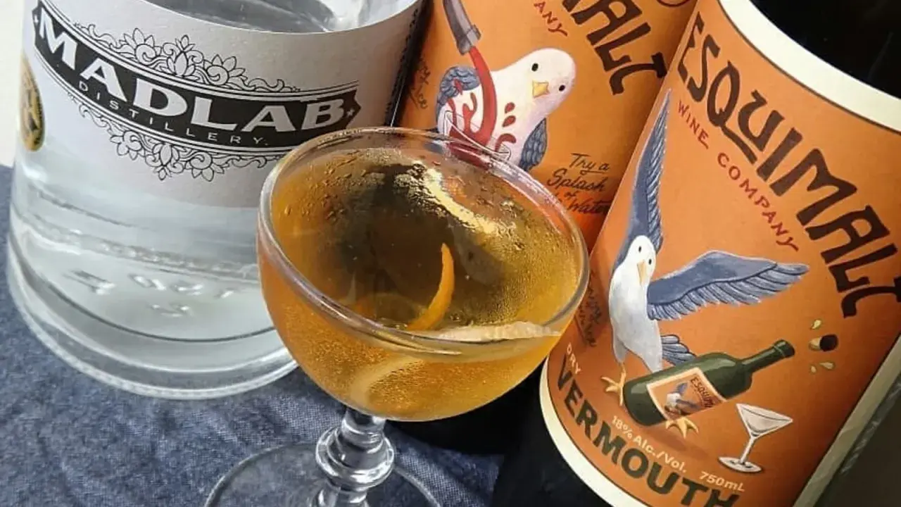 Un cocktail martini doré, garni d'un zeste d'orange, est prêt à être dégusté, entouré de bouteilles de gin et de vermouth.