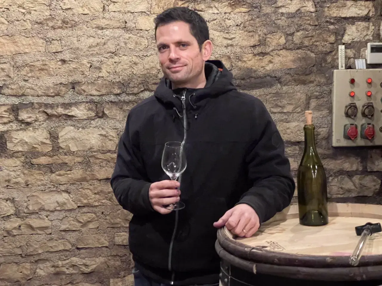 Un homme tient un verre vide dans une cave à vin, à côté d'une bouteille et d'un tonneau. Peut-être une dégustation de Romanée-Conti ?