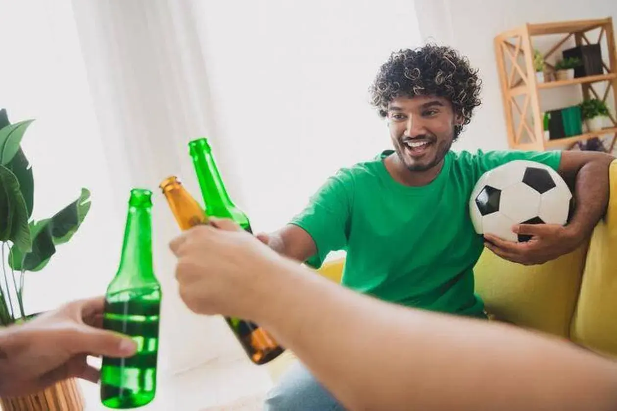 Un homme souriant, ballon de foot sous le bras, trinque avec des bières après le sport.