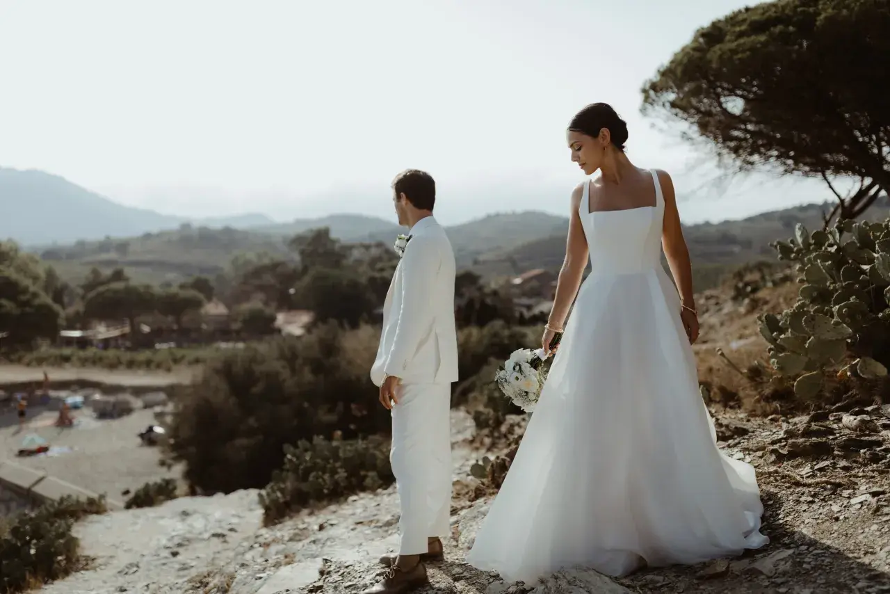 Un couple de mariés en tenue blanche, contemplant le paysage des Clos de Paulilles. La mariée tient un bouquet.